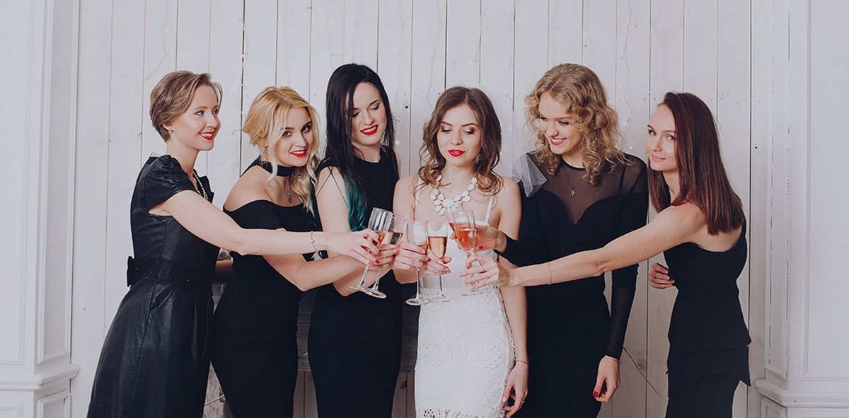 Six women in black dresses and one in white dress toasting. Bridesmaids and bride celebrate with champagne.