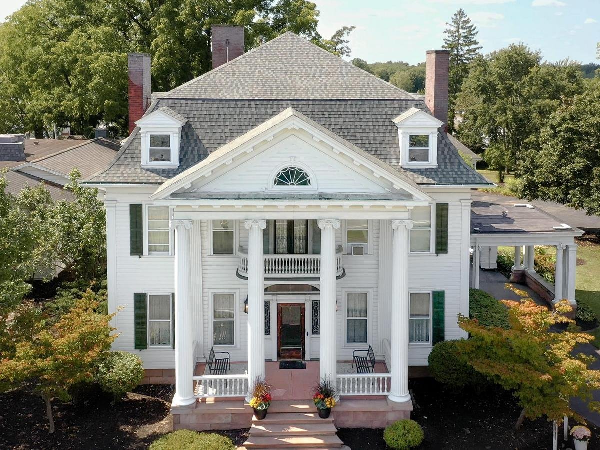 A large white house with columns and green shutters. The house has a gray roof and a well-manicured lawn.