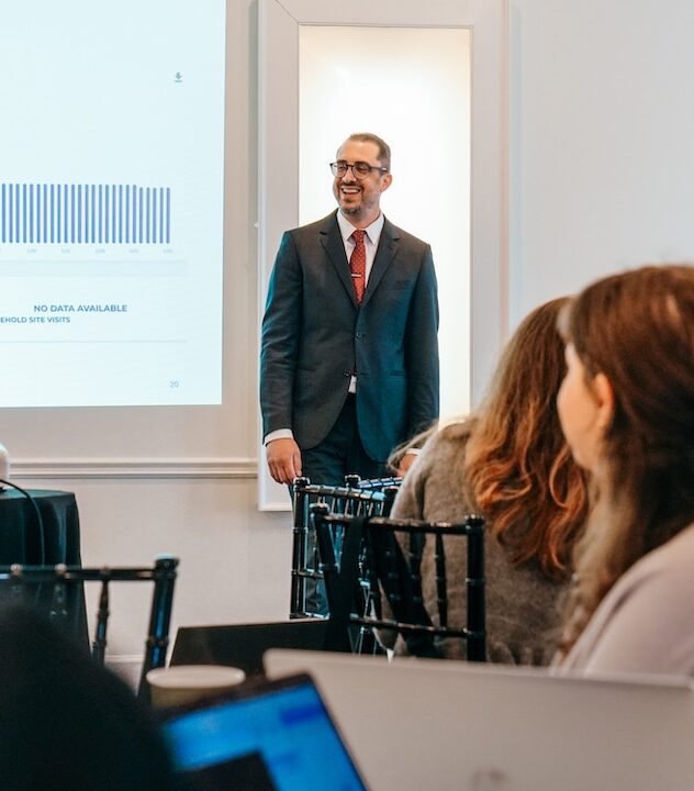 A man in a suit stands before a projector screen. People sit at desks in front of him.