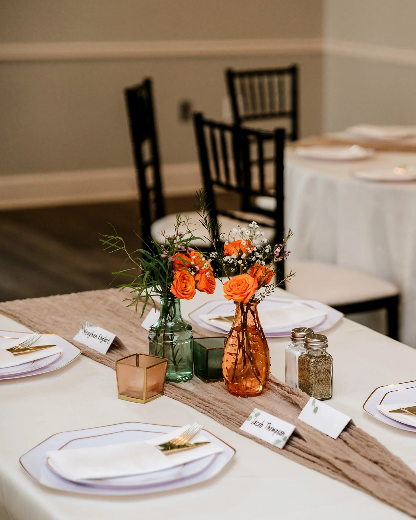 A beautifully set table with white plates, orange flowers, and candles. The table is decorated with a beige runner.