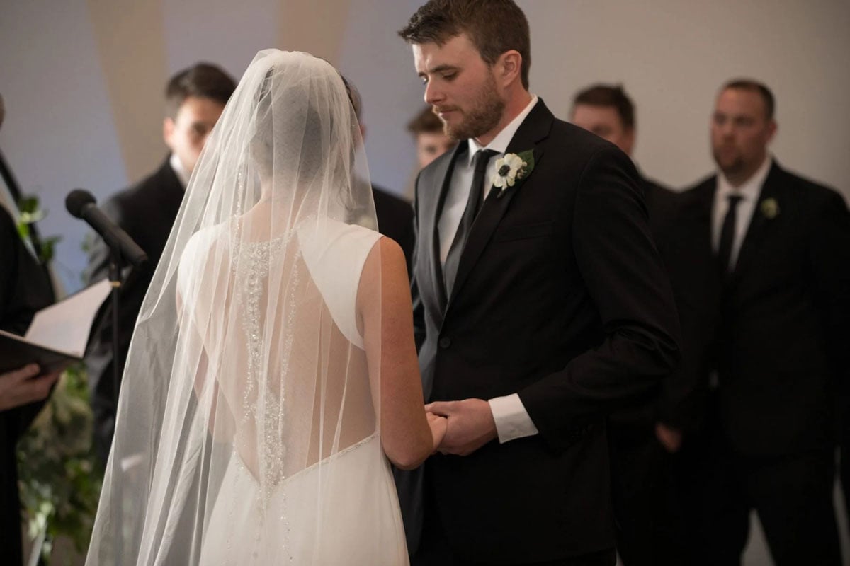 Bride in white dress and veil, groom in black suit. They are holding hands.