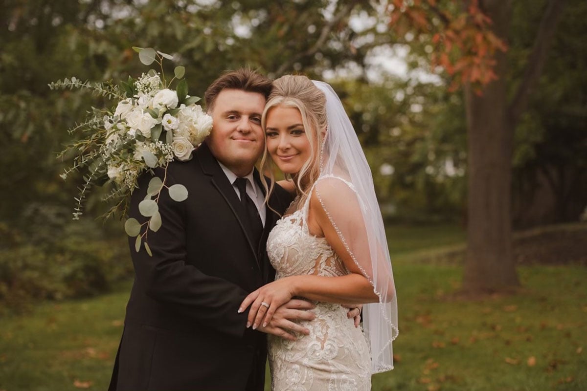 A bride and groom stand together in a grassy area with trees behind them. The bride wears a white dress and veil, and the groom is in a black suit.