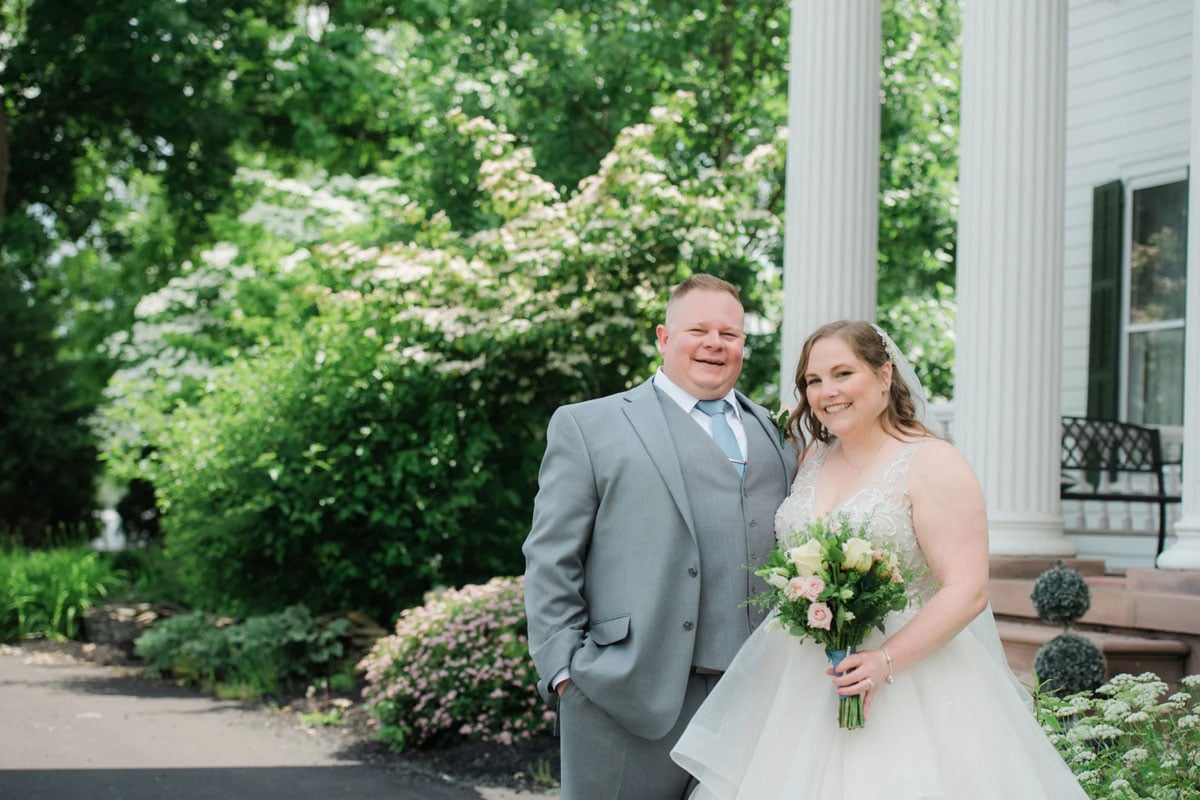 A bride and groom smiling and posing together outside a wedding venue. The bride is holding a bouquet of flowers.