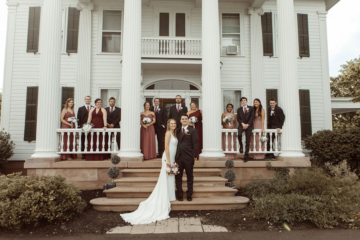 Bride and groom standing on steps with wedding party. The bride wears a white dress and the groom is in a black suit.