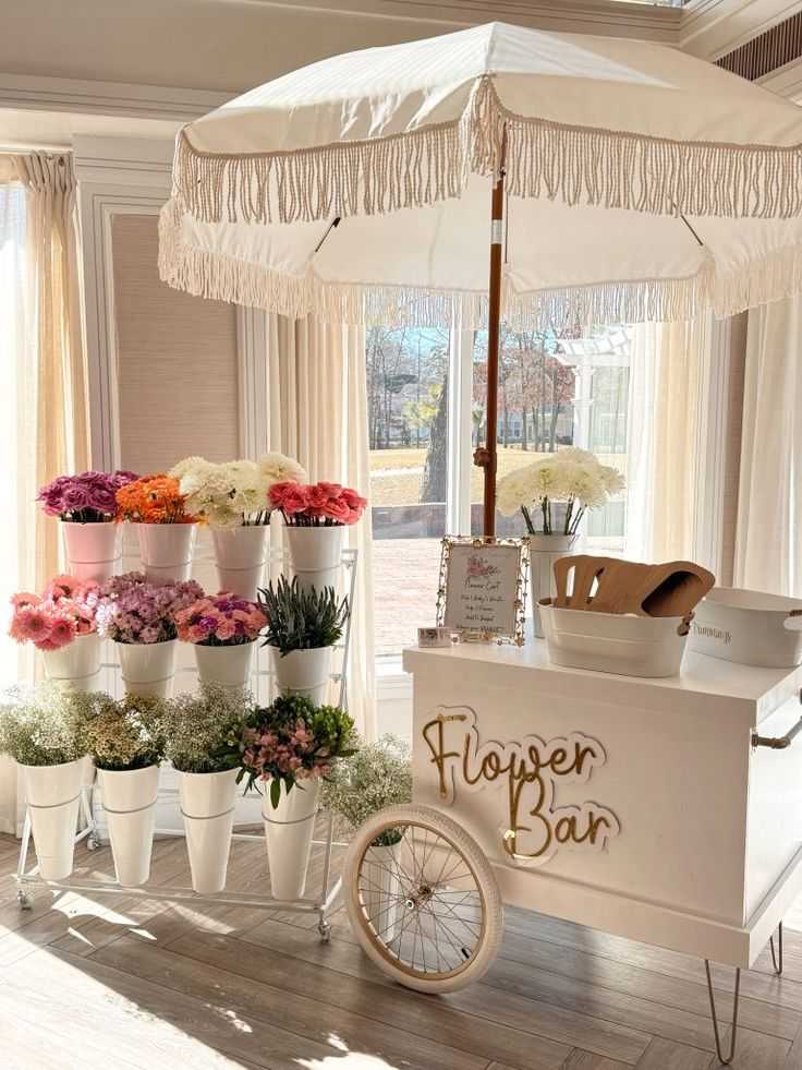 A flower bar with a white umbrella and many potted flowers. The cart reads 'Flower Bar'.