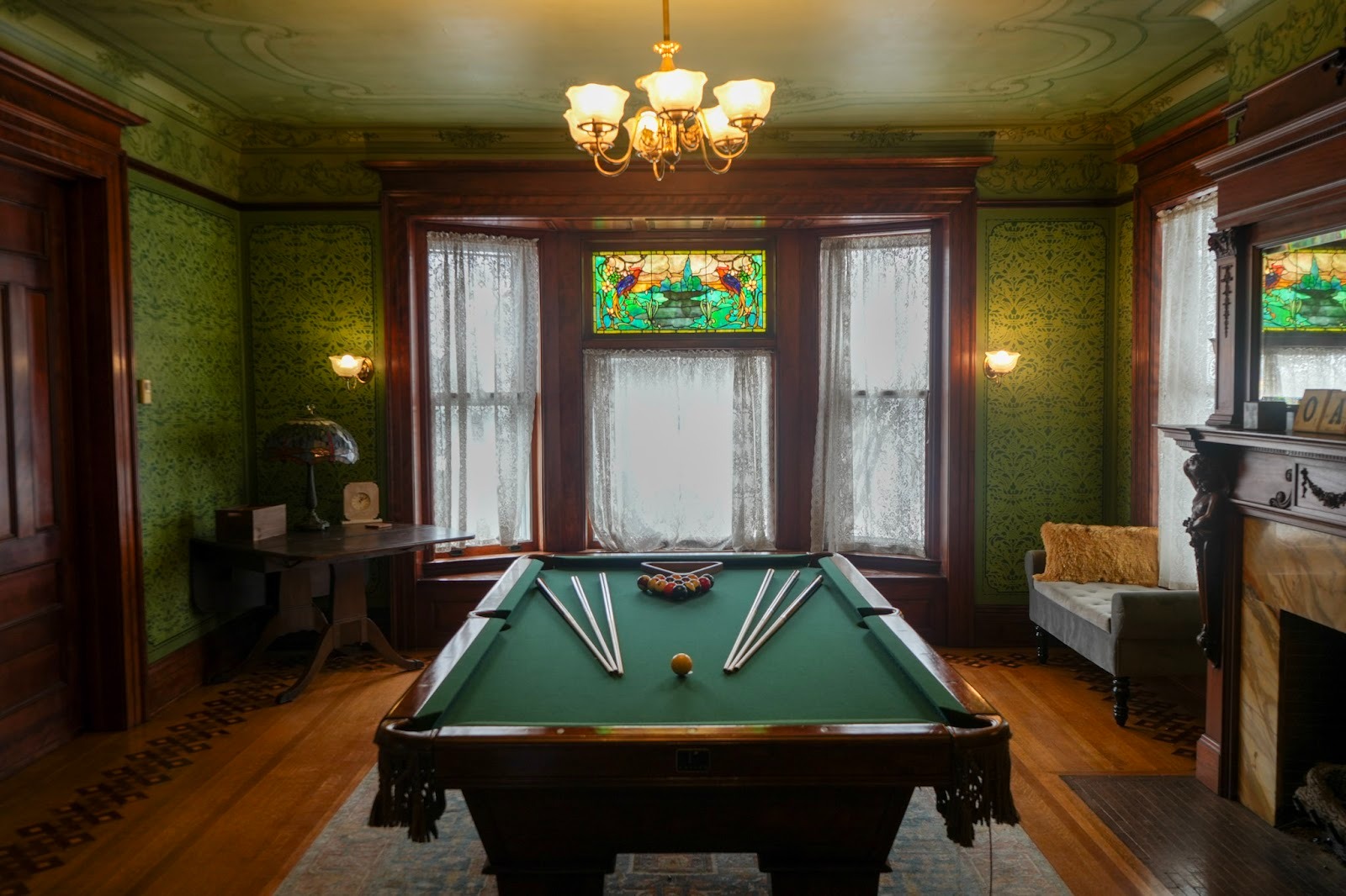 A classic billiard room with green walls and a wooden pool table. The room features large windows with stained glass.