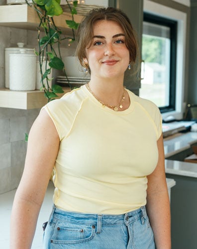 Young woman smiling in a kitchen. She is wearing a yellow top and blue jeans.