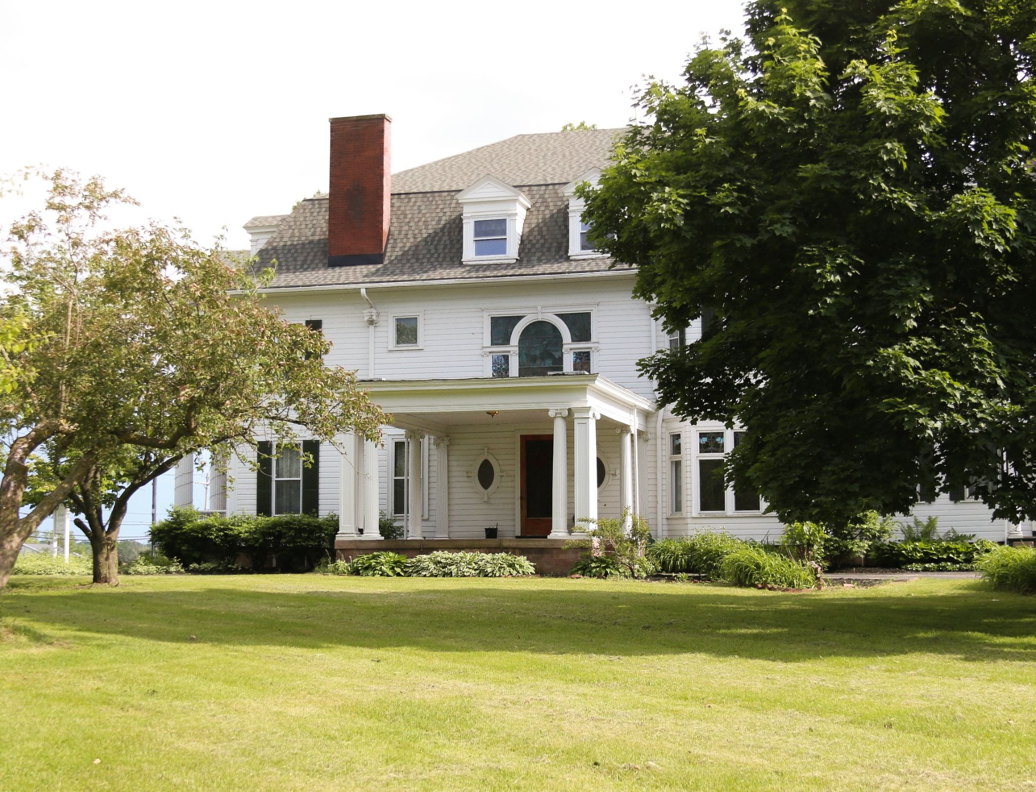 A large white house with a green lawn and trees. The house has a covered porch and a red brick chimney.