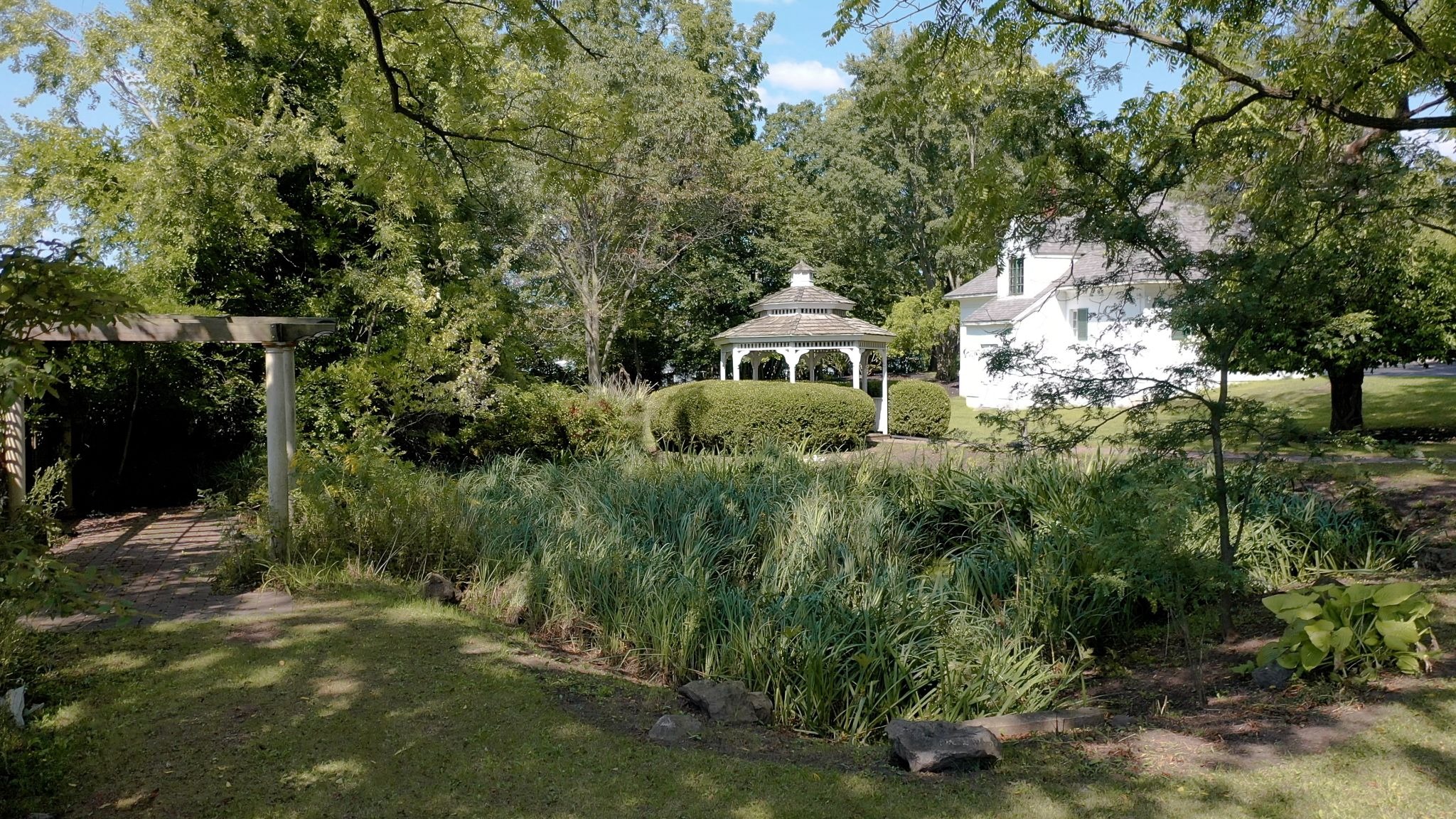A lush garden with a gazebo and a white house in the background. The garden is filled with greenery and trees.