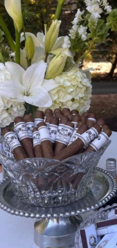 A bowl of cigars on a silver stand with white flowers. The cigars are neatly arranged.