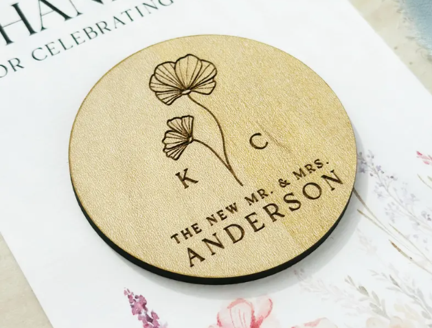 A wooden magnet with a floral design and engraved text. The magnet reads 'K C THE NEW MR. & MRS. ANDERSON'.