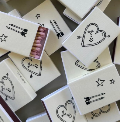 A collection of white matchboxes with cute designs. The boxes have illustrations of hearts, stars, and arrows.