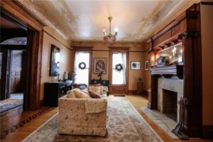 A cozy living room featuring a large ornate fireplace, hardwood floors, and a chandelier. The room includes patterned furniture, decorative wreaths, and a large area rug.