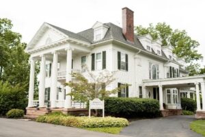 A beautifully landscaped Victorian-style home featuring white columns, dark shutters, and a red brick chimney.