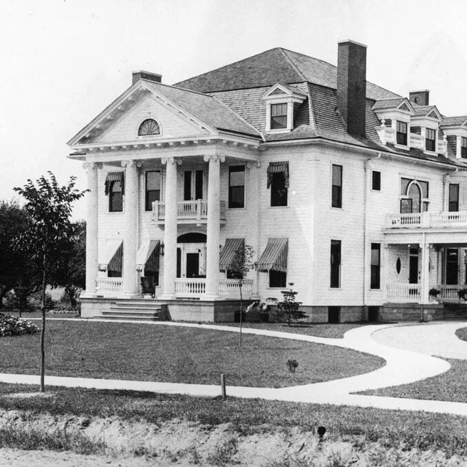A large white mansion with columns and a chimney. The building has multiple stories and a large front yard.