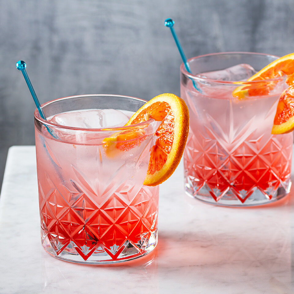 Two glasses of pink cocktail with blue straws and orange slices on a white table.