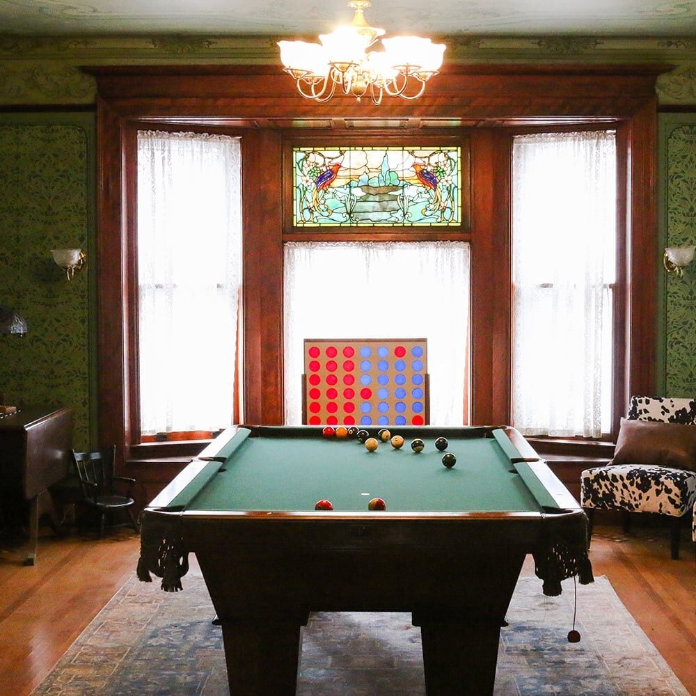 A classic game room with a pool table and stained glass window. The room features green wallpaper and a chandelier.