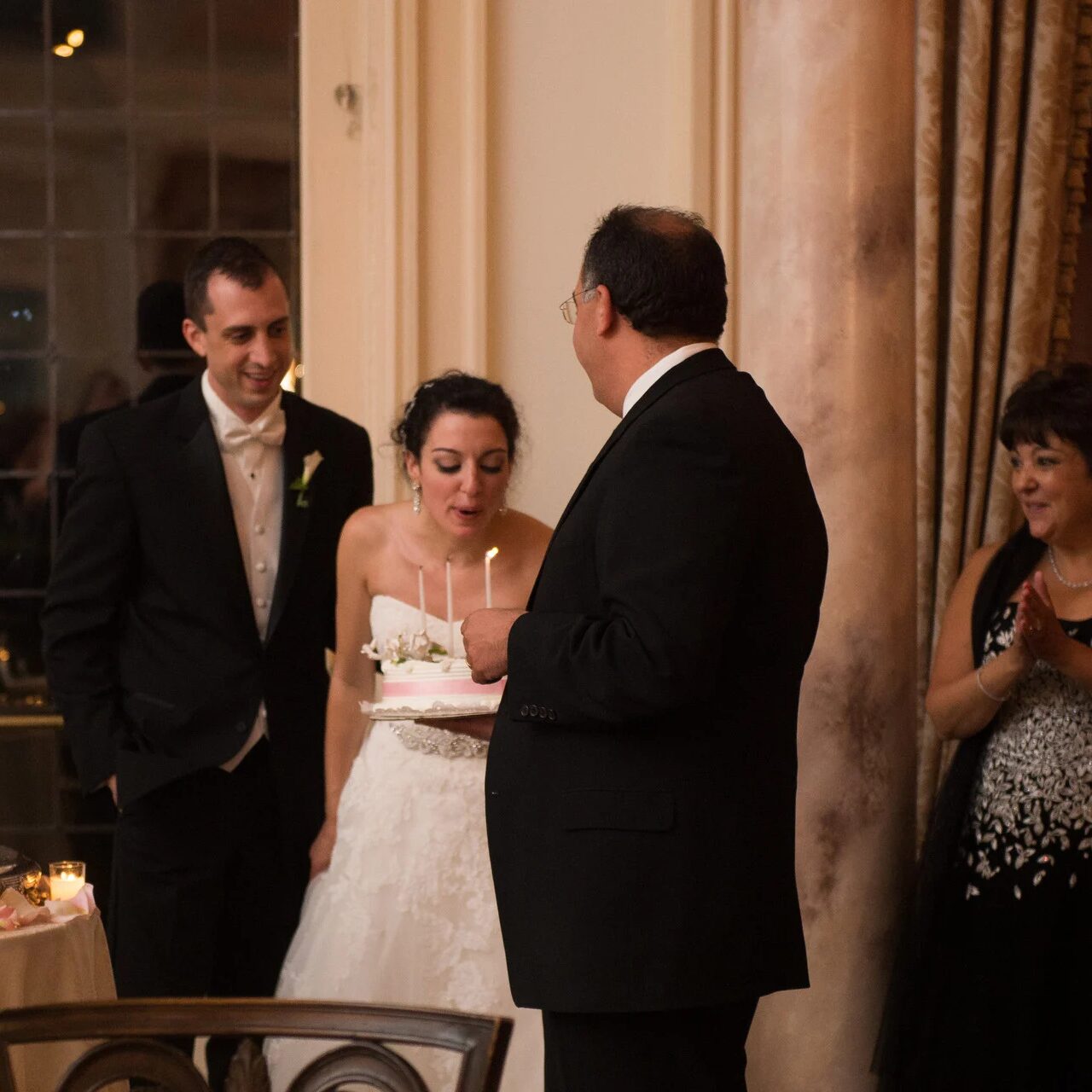 Bride and groom laughing while cutting cake. Wedding party looks on.